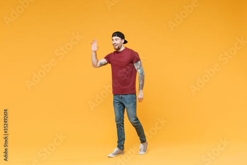 Excited young tattooed man guy in casual t-shirt black cap posing isolated on yellow background studio. People lifestyle concept. Mock up copy space. Waving and greeting with hand as notices someone.