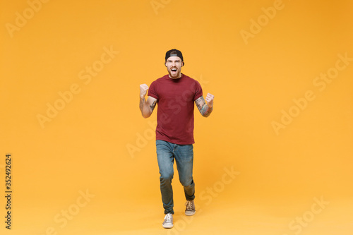 Happy young bearded tattooed man guy in casual t-shirt black cap posing isolated on yellow background in studio. People emotions lifestyle concept. Mock up copy space. Clenching fists like winner.