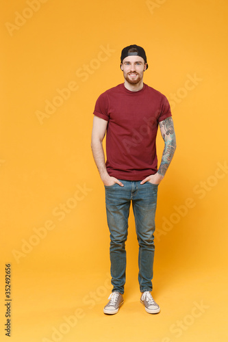 Smiling young bearded tattooed man guy in casual t-shirt black cap posing isolated on yellow wall background studio portrait. People lifestyle concept. Mock up copy space. Holding hands in pockets.