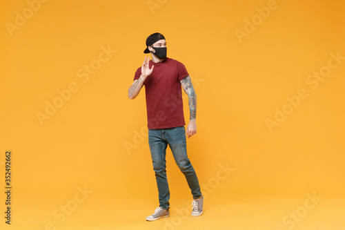 Young tattooed man guy in casual t-shirt cap black face mask posing isolated on yellow background studio. People lifestyle concept. Mock up copy space. Waving greeting with hand as notices someone.