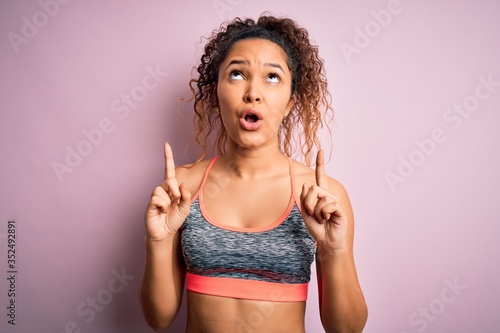 Beautiful sportswoman with curly hair doing sport wearing sportswear over pink background amazed and surprised looking up and pointing with fingers and raised arms. © Krakenimages.com