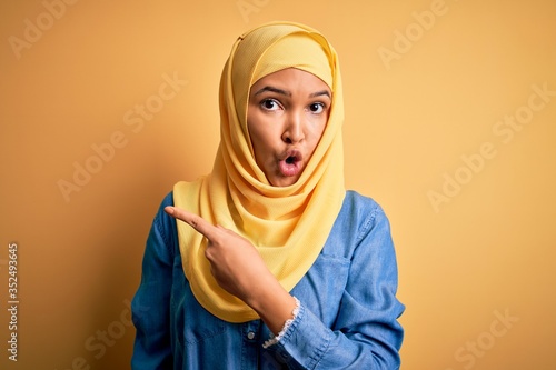 Young beautiful woman with curly hair wearing arab traditional hijab over yellow background Surprised pointing with finger to the side, open mouth amazed expression. © Krakenimages.com