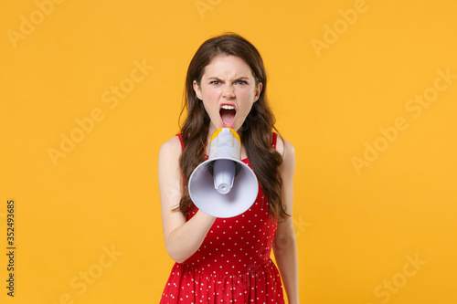 Displeased young brunette woman girl in red summer dress posing isolated on yellow wall background studio portrait. People sincere emotions lifestyle concept. Mock up copy space. Scream in megaphone.