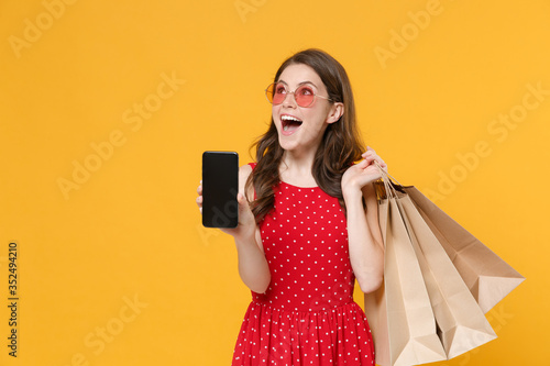 Excited woman girl in red summer dress, eyeglasses isolated on yellow background. People lifestyle concept. Hold package bag with purchases after shopping, hold mobile phone with blank empty screen.
