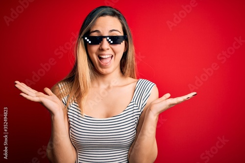 Young beautiful brunette woman wearing funny thug life sunglasses over red background celebrating crazy and amazed for success with arms raised and open eyes screaming excited. Winner concept © Krakenimages.com