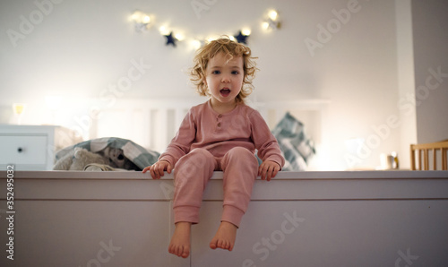 Small toddler girl sitting on bed indoors at home, looking at camera.