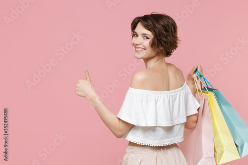 Back rear view of smiling woman girl in summer clothes hold package bag with purchases isolated on pink background. Shopping discount sale concept. Mock up copy space. Showing thumb up looking camera.