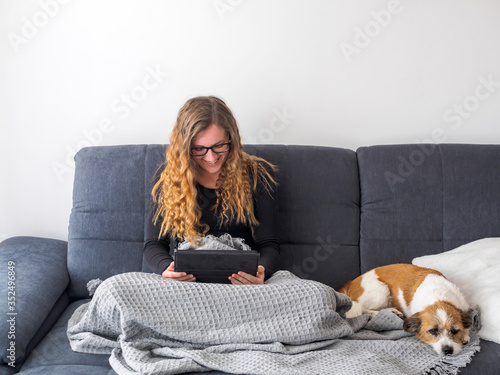 Junge Frau liest in einem Tablett auf ihren Schoß auf einer grauen Couch und daneben schläft ein kleiner Terrier Hund. Hygge, stay home.
