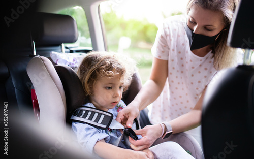 Mother putting small daughter in car seat before trip, wearing face masks.