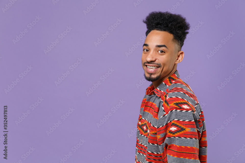Side view of smiling young african american guy in casual colorful shirt posing isolated on violet background studio portrait. People emotions lifestyle concept. Mock up copy space. Looking camera. Side view of smiling young african american guy in casual colorful shirt posing isolated on violet background studio portrait. People emotions lifestyle concept. Mock up copy space. Looking camera.