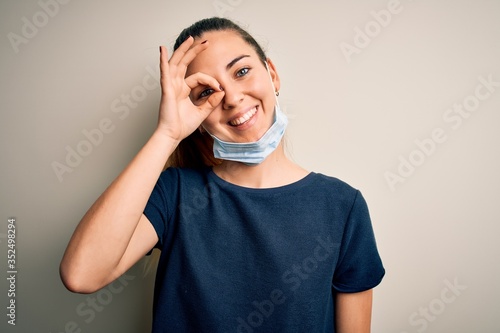 Beautiful blonde woman with blue eyes wearing medical mask over white background doing ok gesture with hand smiling, eye looking through fingers with happy face.