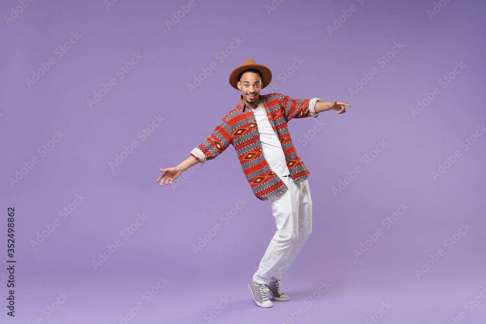 Smiling young african american guy in casual colorful shirt hat posing isolated on violet background studio. People lifestyle concept. Mock up copy space. Dancing, standing on toes, spreading hands. Smiling young african american guy in casual colorful shirt hat posing isolated on violet background studio. People lifestyle concept. Mock up copy space. Dancing, standing on toes, spreading hands.