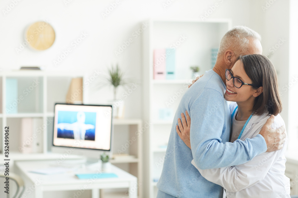 Waist up portrait of young female doctor hugging grateful senior patient and smiling happily while standing in office interior, copy space Waist up portrait of young female doctor hugging grateful senior patient and smiling happily while standing in office interior, copy space