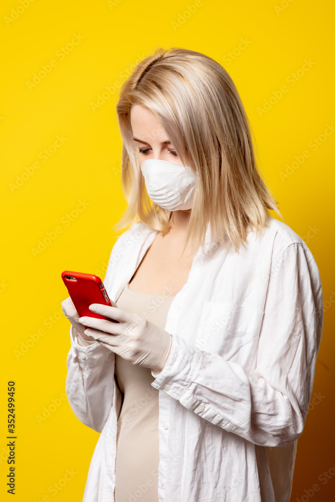 Blonde girl in face mask holding mobile phone on yellow background. Blonde girl in face mask holding mobile phone on yellow background.