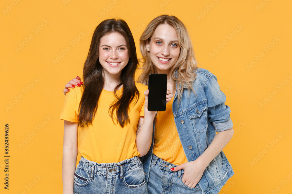Two smiling young women girls friends in casual t-shirts denim clothes posing isolated on yellow wall background studio portrait. People lifestyle concept. Hold mobile phone with blank empty screen. Two smiling young women girls friends in casual t-shirts denim clothes posing isolated on yellow wall background studio portrait. People lifestyle concept. Hold mobile phone with blank empty screen.