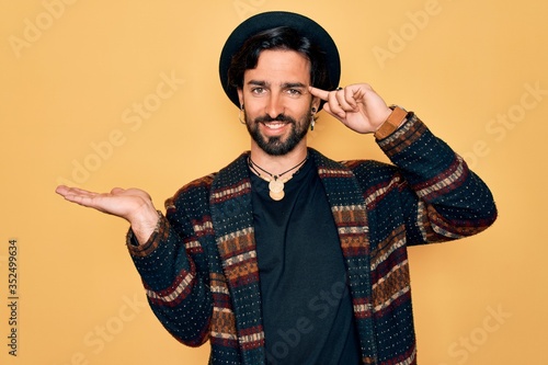 Young handsome hispanic bohemian man wearing hippie style and boho hat confused and annoyed with open palm showing copy space and pointing finger to forehead. Think about it.
