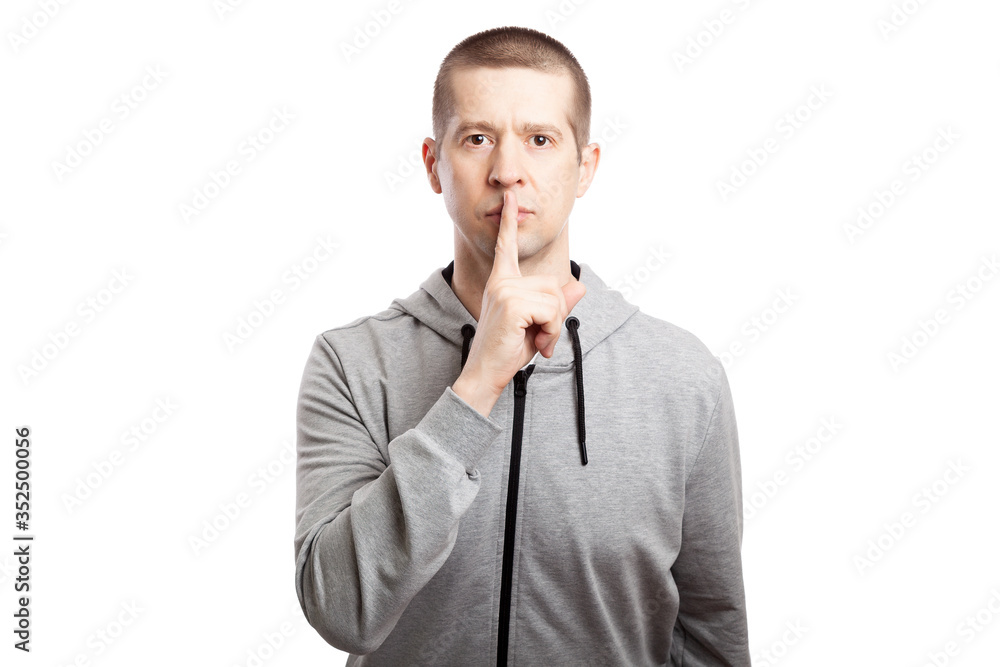healthy, athletic male athlete shows a gesture of silence and mystery, asks to remain silent, puts his index finger to his lips. Dressed in a grey hooded jacket. A look into the camera. Isolated healthy, athletic male athlete shows a gesture of silence and mystery, asks to remain silent, puts his index finger to his lips. Dressed in a grey hooded jacket. A look into the camera. Isolated