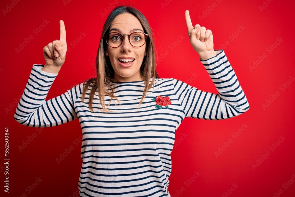 Young beautiful blonde woman with blue eyes wearing glasses standing over red background smiling amazed and surprised and pointing up with fingers and raised arms. Young beautiful blonde woman with blue eyes wearing glasses standing over red background smiling amazed and surprised and pointing up with fingers and raised arms.