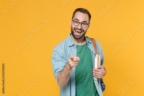 Cheerful young man student in casual clothes glasses with backpack hold books isolated on yellow background studio. Education in high school university college concept. Point index finger on camera.