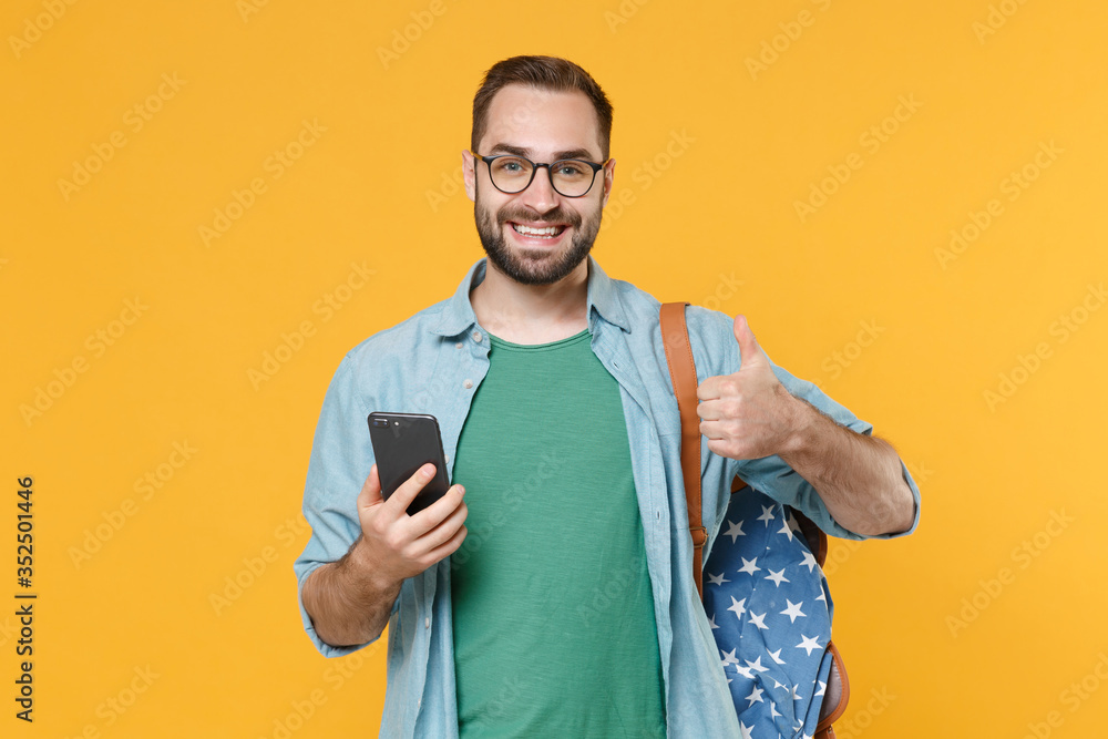 Smiling young man student in casual clothes glasses with backpack posing isolated on yellow background studio. Education in high school university college concept. Using mobile phone showing thumb up. Smiling young man student in casual clothes glasses with backpack posing isolated on yellow background studio. Education in high school university college concept. Using mobile phone showing thumb up.