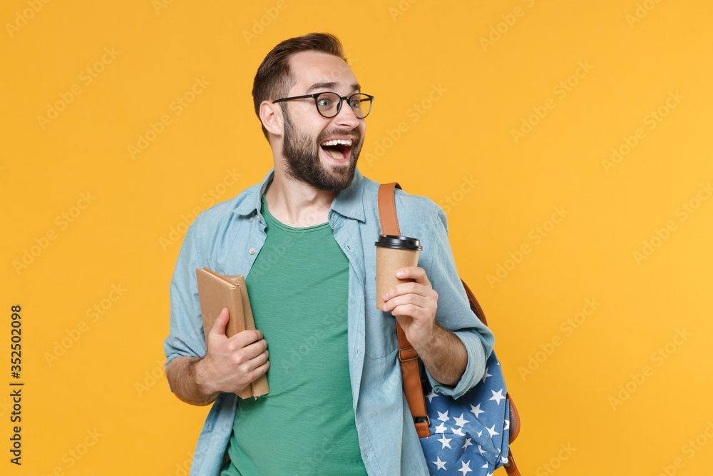 Amazed man student in glasses backpack hold books isolated on yellow background. Education in high school university college concept. Mock up copy space. Hold paper cup of coffee or tea looking aside. Amazed man student in glasses backpack hold books isolated on yellow background. Education in high school university college concept. Mock up copy space. Hold paper cup of coffee or tea looking aside.