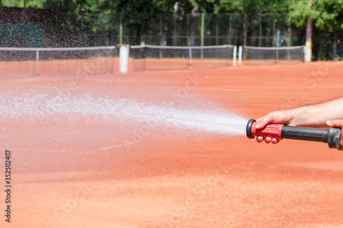 Hose sprinkler in the hand of a worker, jet and spray of water and blurry clay tennis courts background. Maintenance of clay tennis court. Service and preparation of the court. Close up. Copy space.