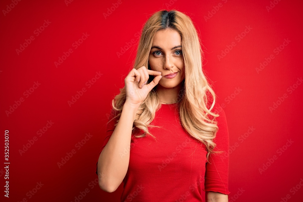 Young beautiful blonde woman wearing casual t-shirt standing over isolated red background mouth and lips shut as zip with fingers. Secret and silent, taboo talking Young beautiful blonde woman wearing casual t-shirt standing over isolated red background mouth and lips shut as zip with fingers. Secret and silent, taboo talking