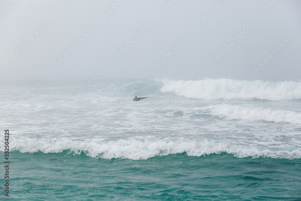Extreme young surfboarder paddling to line-up in foggy weather with low visibility. Lonely surfer in the blue ocean waves. Surfing concept