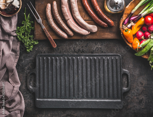 Grill or barbecue food background. Empty cast iron grill griddle and meat fork on rustic kitchen table. Various sausages on a wooden cutting board. Wooden bowl with vegetables. Top view.