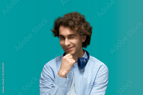 Blink. Portrait of a young beautiful man wearing white t-shirt and blue shirt in blue headphones.Thought and holds a fist at the chin and wink at camera