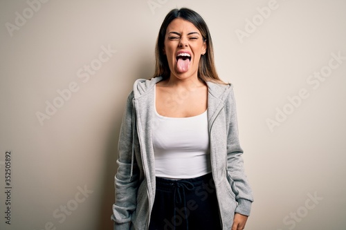 Young beautiful brunette sportswoman wearing sportswoman training over white background sticking tongue out happy with funny expression. Emotion concept.