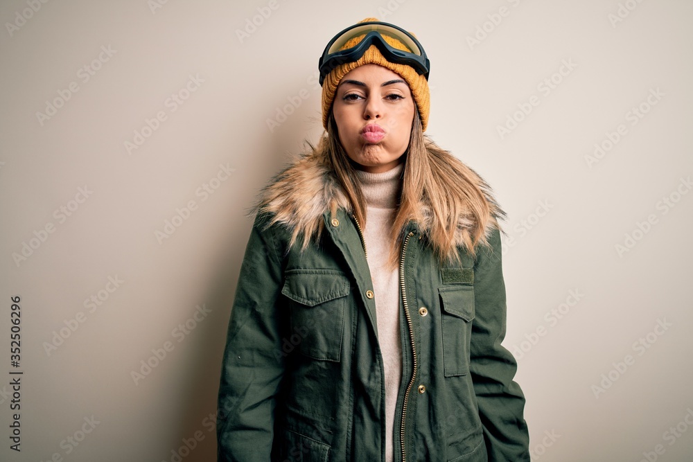 Young brunette skier woman wearing snow clothes and ski goggles over white background puffing cheeks with funny face. Mouth inflated with air, crazy expression.
