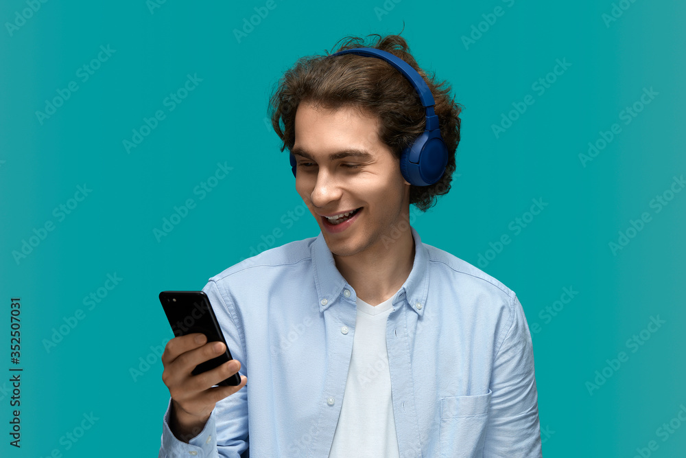 Funny sound. Portrait of a young beautiful man wearing white t-shirt and blue shirt in blue headphones holding mobile phone in hand and looking at him with smile Funny sound. Portrait of a young beautiful man wearing white t-shirt and blue shirt in blue headphones holding mobile phone in hand and looking at him with smile