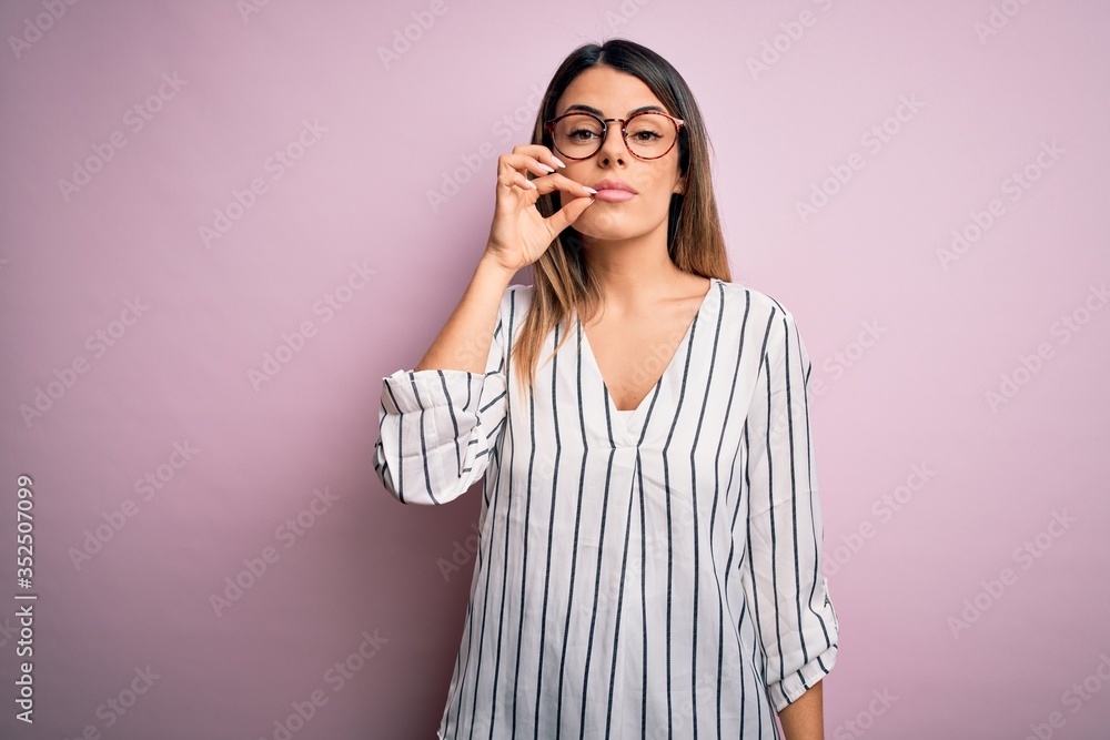 Young beautiful woman wearing casual striped t-shirt and glasses over pink background mouth and lips shut as zip with fingers. Secret and silent, taboo talking Young beautiful woman wearing casual striped t-shirt and glasses over pink background mouth and lips shut as zip with fingers. Secret and silent, taboo talking