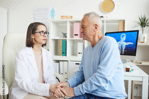 Portrait of caring female doctor holding hands with senior patient while discussing diagnosis during consultation, copy space