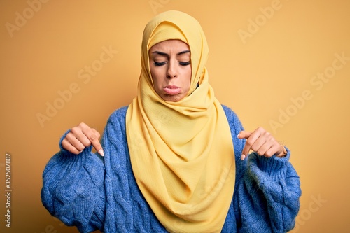 Young beautiful brunette muslim woman wearing arab hijab over isolated yellow background Pointing down looking sad and upset, indicating direction with fingers, unhappy and depressed. © Krakenimages.com