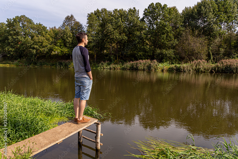A young guy in denim shorts and a longsleeve stands on a wooden structure by the river, with his hands in his pockets and looking at the river, back and side view. A young guy in denim shorts and a longsleeve stands on a wooden structure by the river, with his hands in his pockets and looking at the river, back and side view.