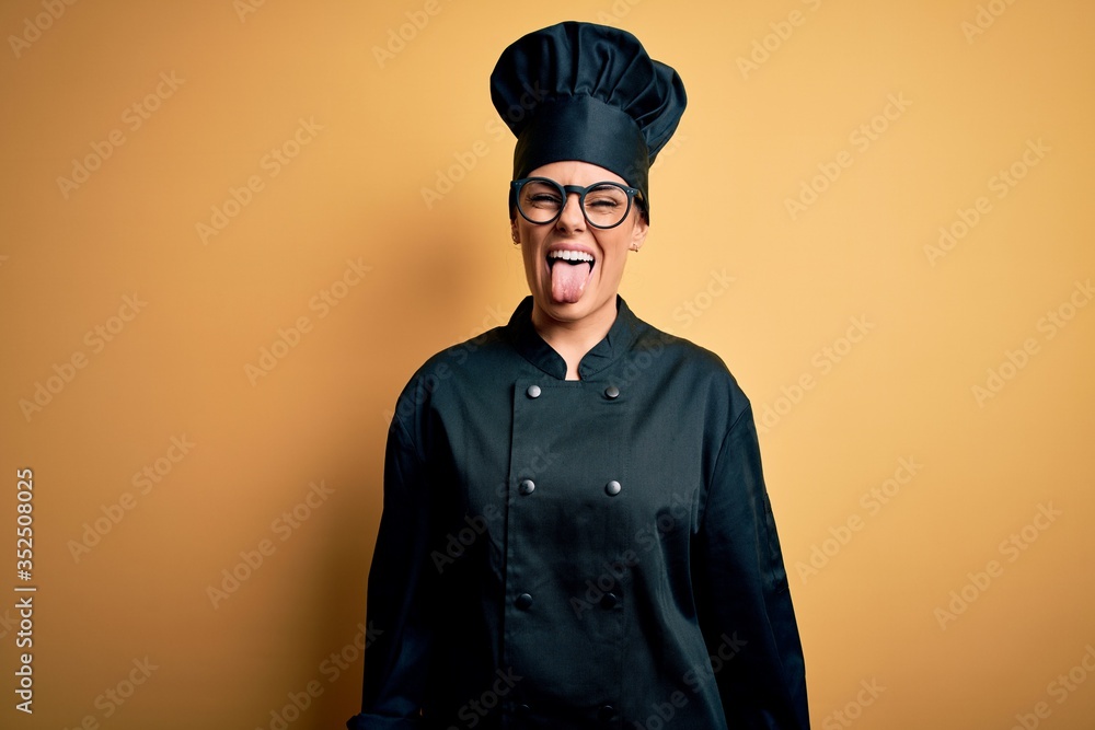 Young beautiful brunette chef woman wearing cooker uniform and hat over yellow background sticking tongue out happy with funny expression. Emotion concept. Young beautiful brunette chef woman wearing cooker uniform and hat over yellow background sticking tongue out happy with funny expression. Emotion concept.