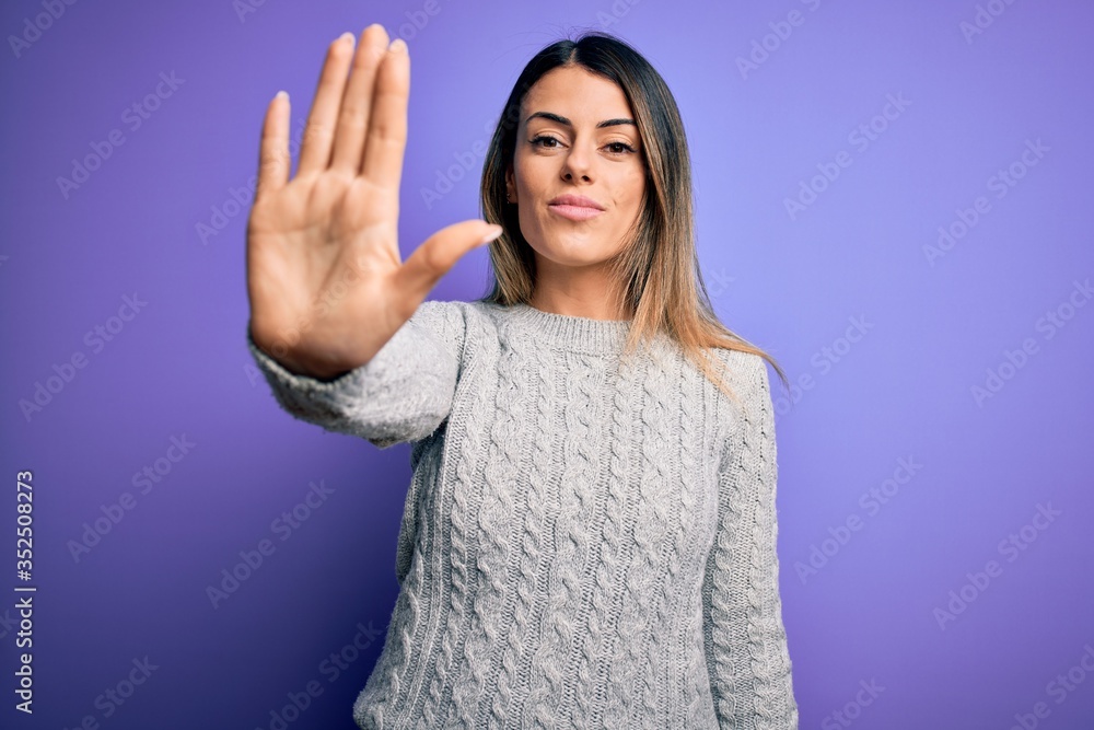 Young beautiful woman wearing casual sweater standing over isolated purple background doing stop sing with palm of the hand. Warning expression with negative and serious gesture on the face. Young beautiful woman wearing casual sweater standing over isolated purple background doing stop sing with palm of the hand. Warning expression with negative and serious gesture on the face.