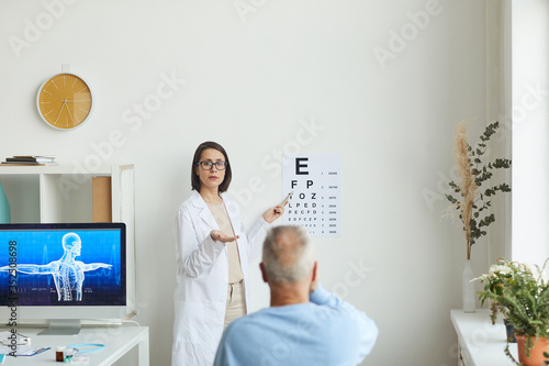 Wide angle portrait of young female ophthalmologist pointing at eye chart while testing eyesight of senior patient, copy space photo