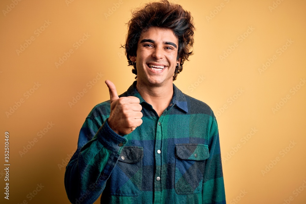 Young handsome man wearing casual shirt standing over isolated yellow background doing happy thumbs up gesture with hand. Approving expression looking at the camera showing success. Young handsome man wearing casual shirt standing over isolated yellow background doing happy thumbs up gesture with hand. Approving expression looking at the camera showing success.