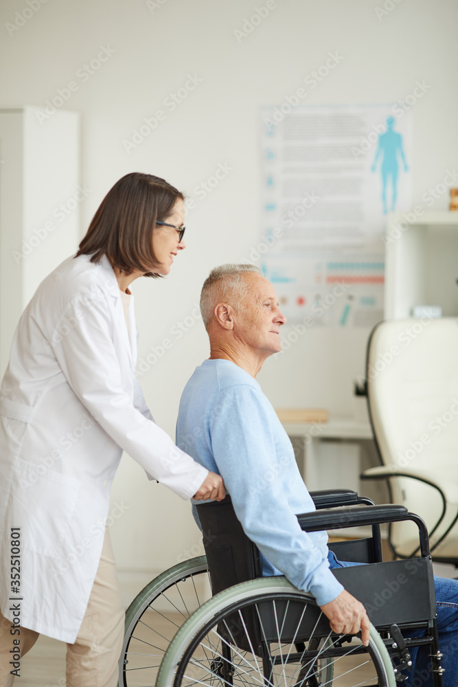Vertical side view portrait of smiling female doctor pushing senior man in wheelchair for examination or consultation in clinic, copy space Vertical side view portrait of smiling female doctor pushing senior man in wheelchair for examination or consultation in clinic, copy space