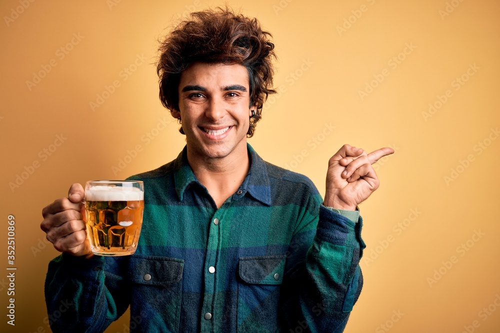 Young handsome man drinking jar of beer standing over isolated yellow background very happy pointing with hand and finger to the side Young handsome man drinking jar of beer standing over isolated yellow background very happy pointing with hand and finger to the side