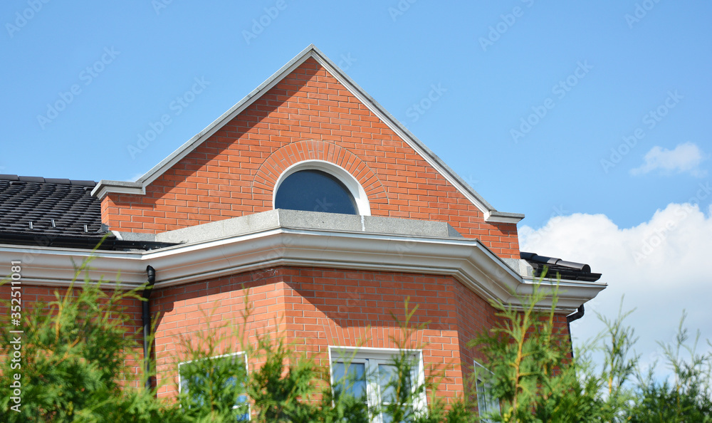 A close-up on a brick house facade with a cornice, decorative molding and attic round window under metal roof.