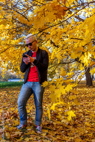 A young short-haired blonde man wearing black jacket texting in the autumn park. Meditation in the falling park.