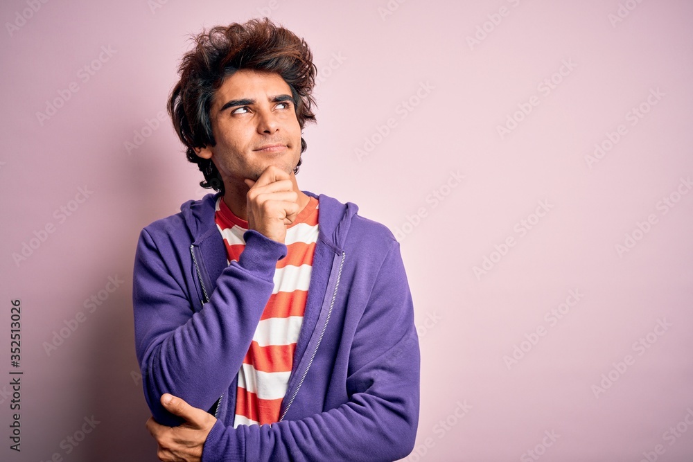 Young man wearing striped t-shirt and purple sweashirt over isolated pink background with hand on chin thinking about question, pensive expression. Smiling with thoughtful face. Doubt concept.