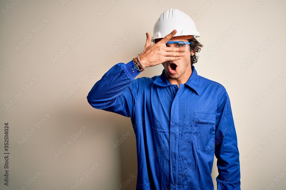 Young constructor man wearing uniform and security helmet over isolated white background peeking in shock covering face and eyes with hand, looking through fingers with embarrassed expression.