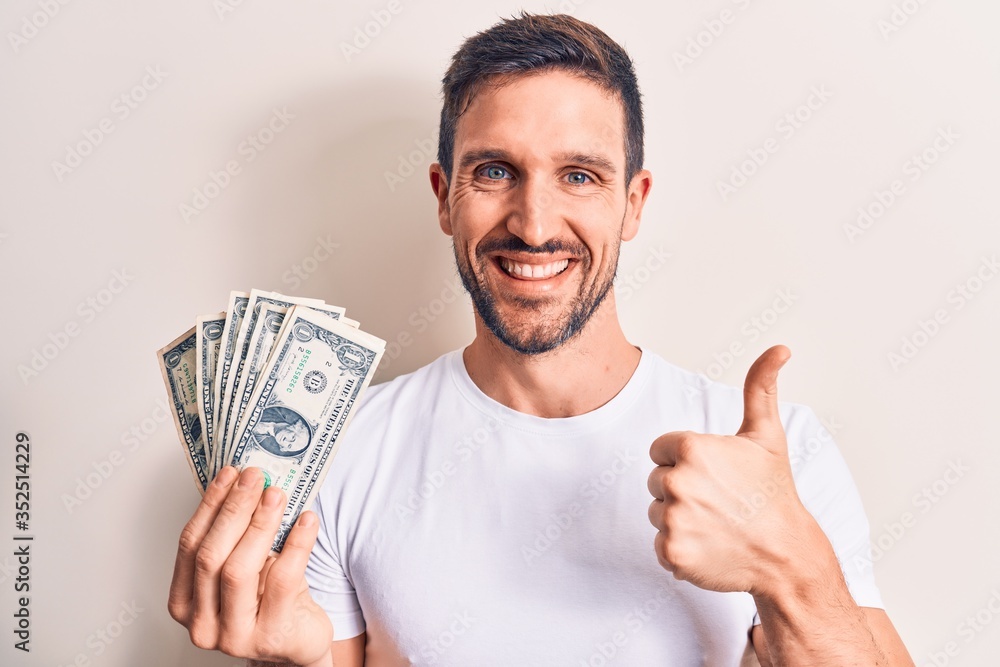Young handsome man holding bunch of dollars banknotes over isolated white background smiling happy and positive, thumb up doing excellent and approval sign Young handsome man holding bunch of dollars banknotes over isolated white background smiling happy and positive, thumb up doing excellent and approval sign