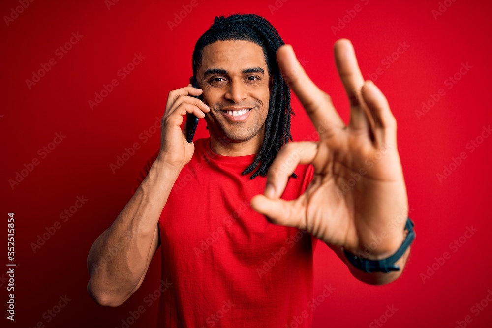 Fototapeta premium Young african american afro man with dreadlocks having conversation talking on smartphone doing ok sign with fingers, excellent symbol