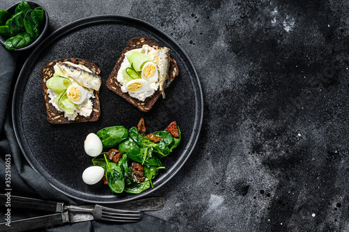 Sandwiches with sardines, egg, cucumber and cream cheese, salad garnish with spinach and dried tomatoes. Black background. Top view. Copy space